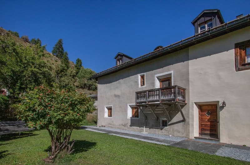 2-story house with balcony, wooden door, well-maintained exterior with gravel pathways, and lush green surroundings.