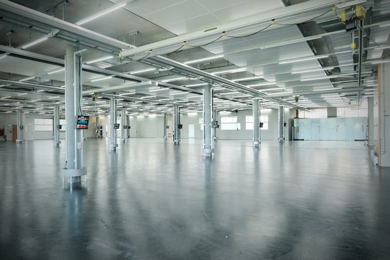 Warehouse or factory space with metal support beams, high ceilings, grey flooring, fluorescent lights, large doors, industrial monitors, and multiple electrical cords