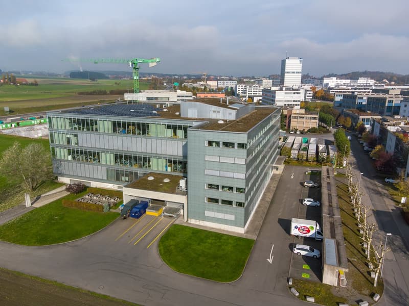 Office building, 4 stories, large windows, grey and white exterior, solar panels on the roof, parking area with cars