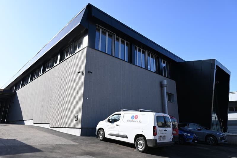 Modern industrial building, gray exterior, multiple windows, solar panels on the roof, cars parked in front