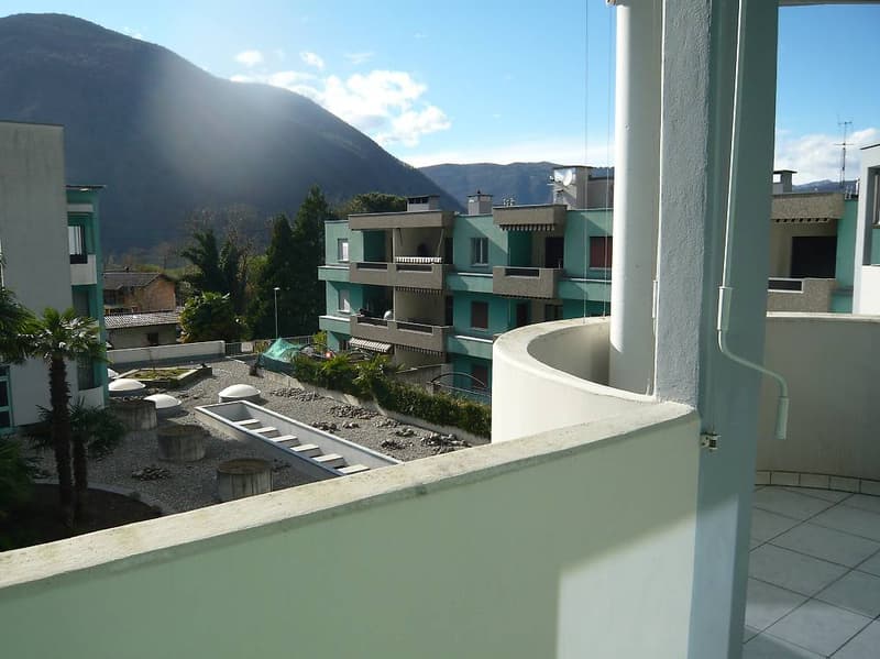 A balcony with a view of a mountain, a building, and a garden with a concrete pathway and a green tent.