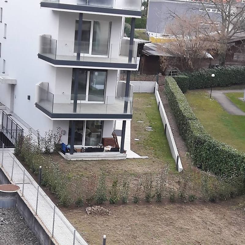 Modern apartment building, white exterior, glass balconies, multiple windows, small front garden, low bushes, fence, path, street light