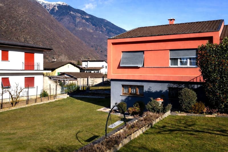 House with orange facade, brown roof, blue lower part, garden in front with lawn and plants, mountains in the background