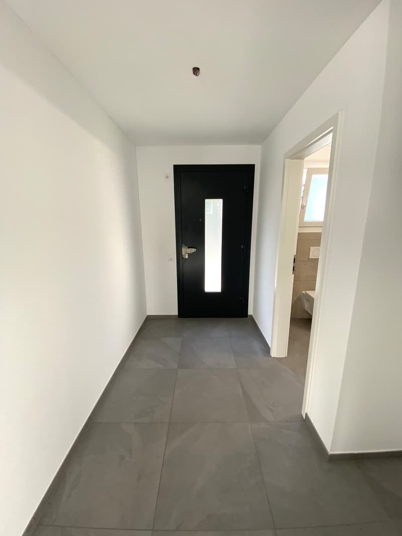 Empty room with white walls and tile flooring. There is a black door, and an opened door with a bathroom visible.