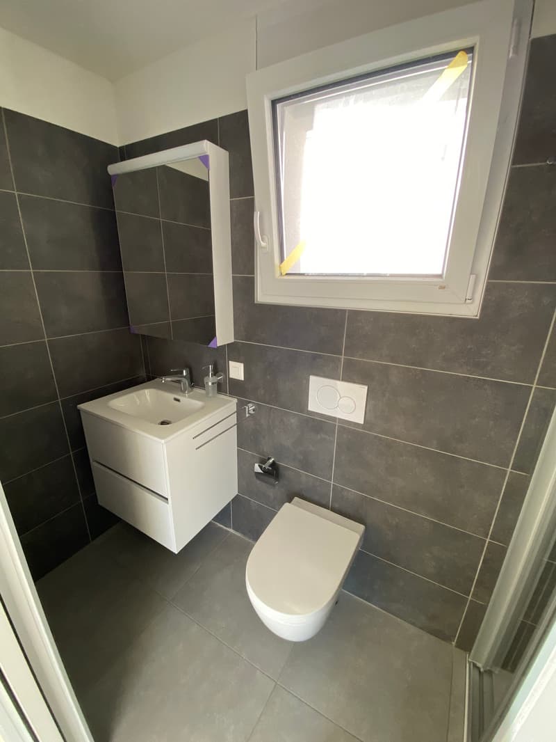 Modern, compact bathroom featuring a white sink with cabinets, a white toilet, and a gray tile floor and walls with a glass window
