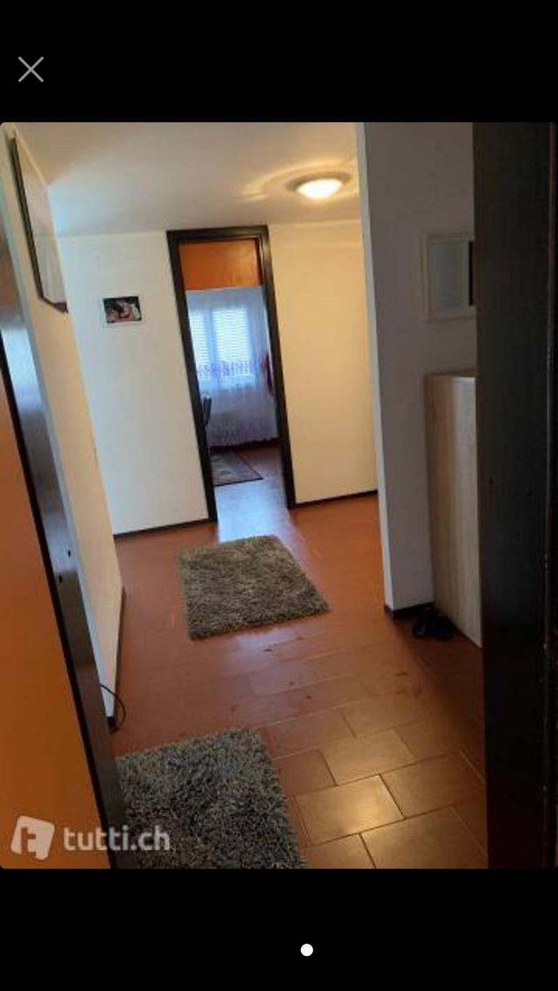 Empty hallway with wooden floor, two rugs, open door, white walls, ceiling light, picture frames, and a white cabinet.