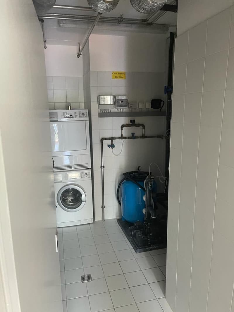 A small, empty utility room with white tiled flooring and walls. It contains a stack of a washing machine and a dryer, and a water tank with a pump system. There is a yellow sign on the wall and a metal pipe on the wall.