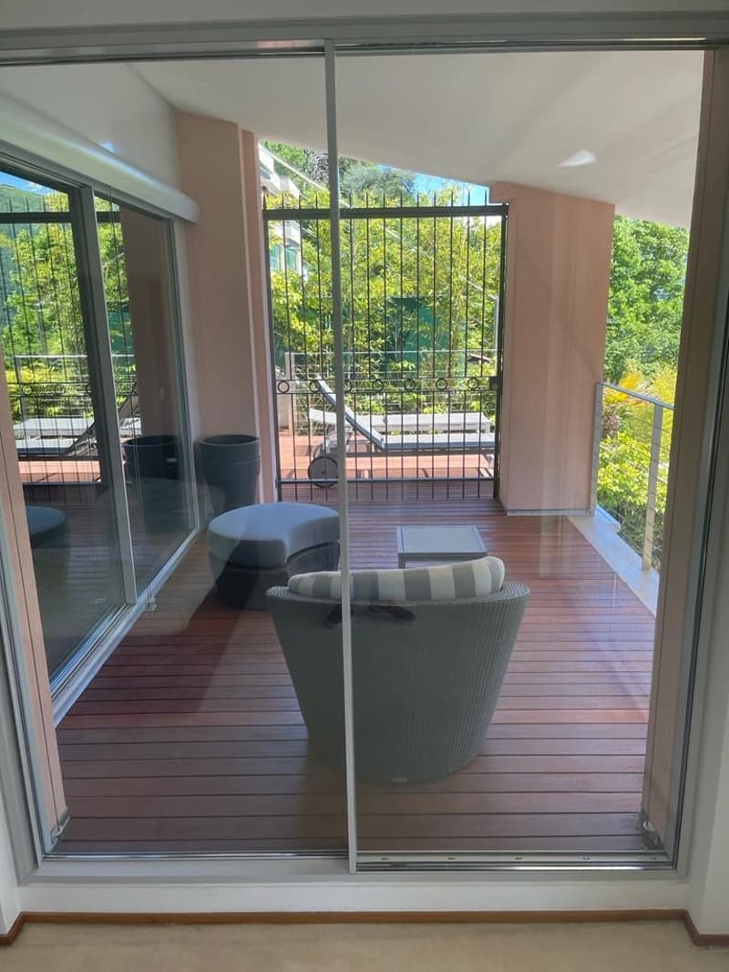 A balcony with a wooden floor, a chair with a cushion, a small table, and a plant pot. There is a bench and a metal railing.