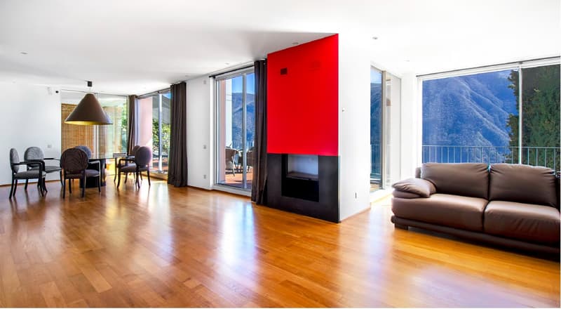 Spacious living room with hardwood floors, large windows, and a red accent wall. Features a dining area with a table and chairs, a fireplace, and a couch. Offers a view of the mountains.