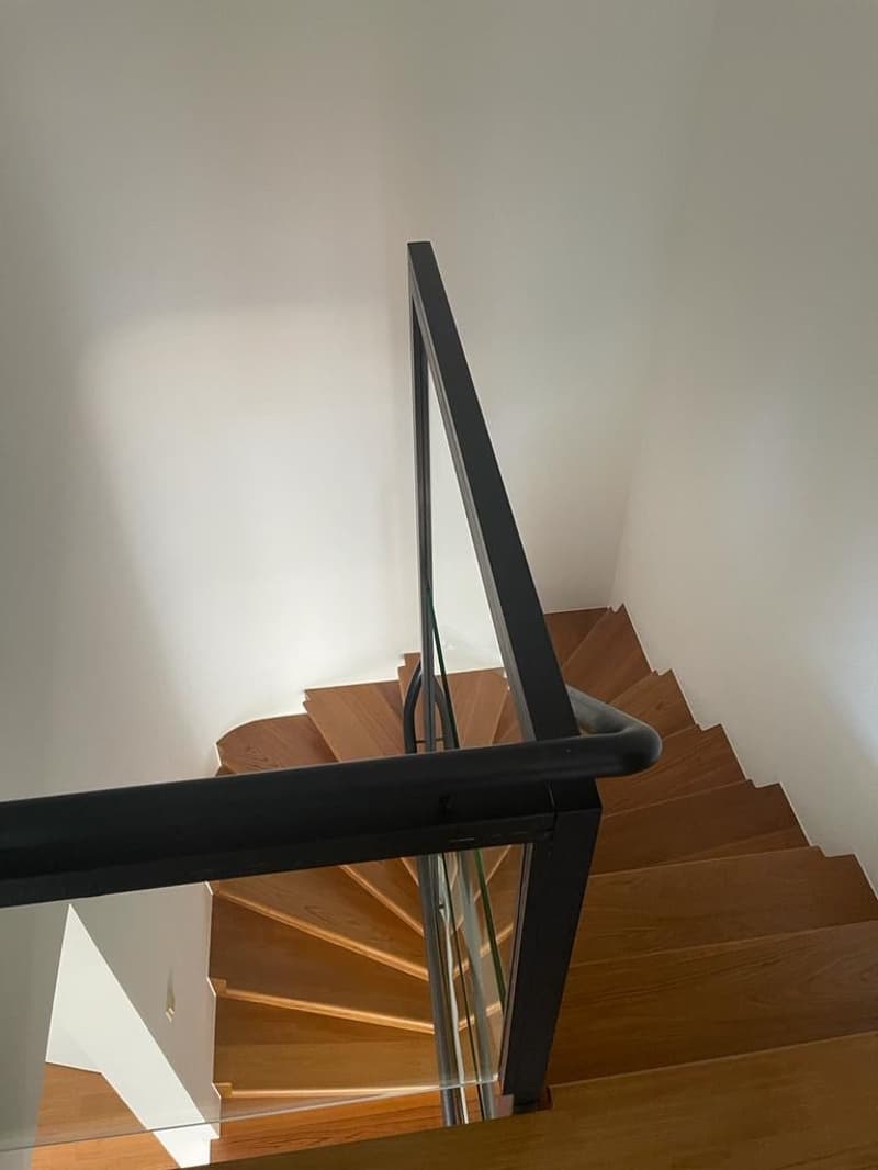 Modern staircase with wooden steps and black metal railing, glass paneling, and white walls.