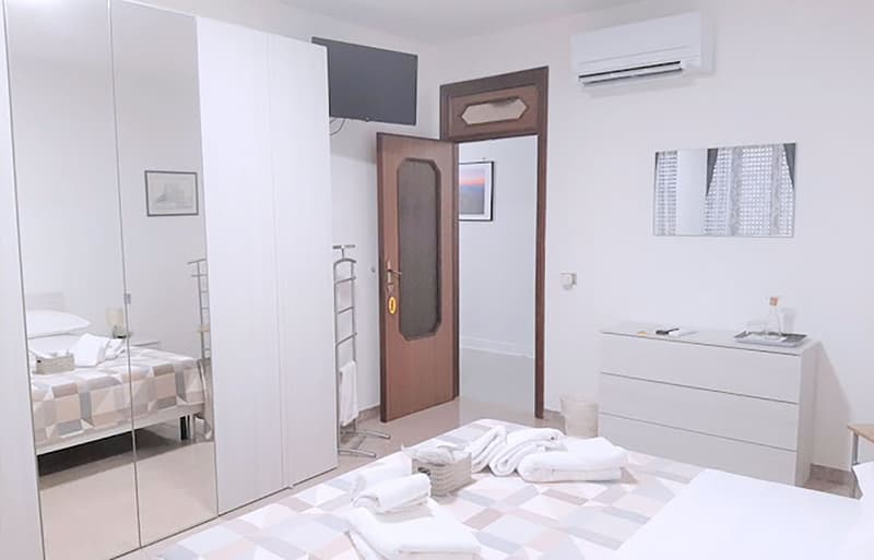 modern interior, bed with white sheets and pillows, mirror wardrobes, TV mounted on wall, wooden floor, air conditioner, and wooden door