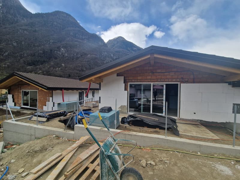 Two houses under construction, one with a brown roof and the other with a black roof, surrounded by a mountainous landscape.