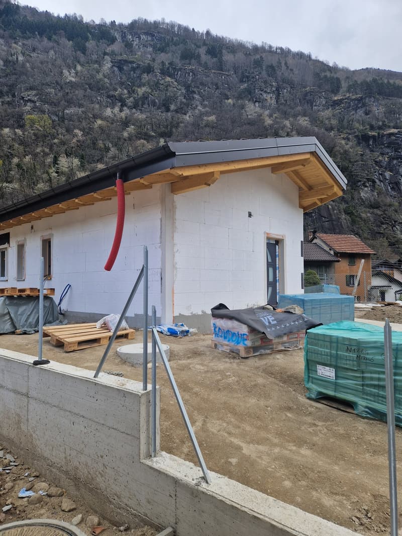 A house under construction with a white exterior, wooden beams, and a gray roof. The house is surrounded by a construction site with dirt, wooden pallets, and metal poles. In the background, there is a mountain with trees and a house.