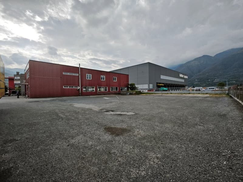 Industrial building with red and gray exterior, multiple windows, large parking lot in front, mountains in the background