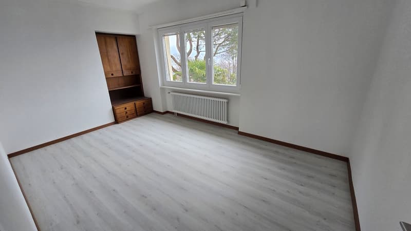 empty room with white walls and flooring, large window, wooden cabinet, white radiator