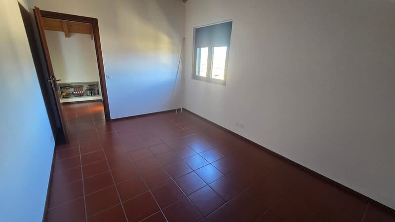 Empty room with white walls, brown tiled floor, small window, and an open door leading to another room