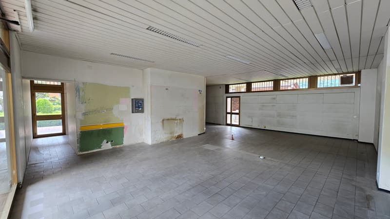 An empty room with tiled flooring, white walls, and a ceiling with white panels and lights. There are two doors on the left side, one of which is open, and a small object on the floor. The room has a few power outlets on the wall and a small piece of pape