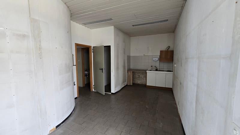 An empty room with white walls and tiled floor. There are two doors, one of which is open, leading to a small bathroom with a sink and a toilet. There is also a small kitchen area with a sink, cabinets, and a countertop.