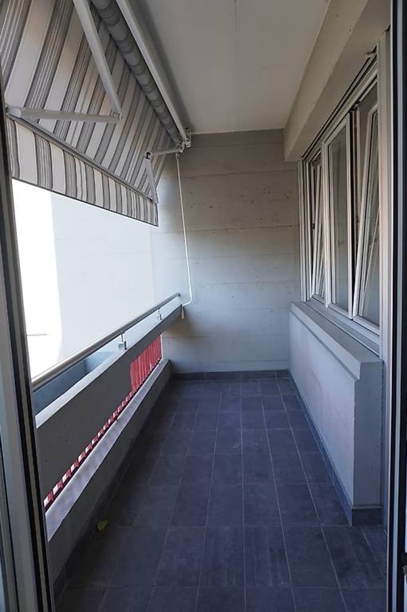 Balcony with grey tile flooring, glass doors, retractable shade, railings, windows