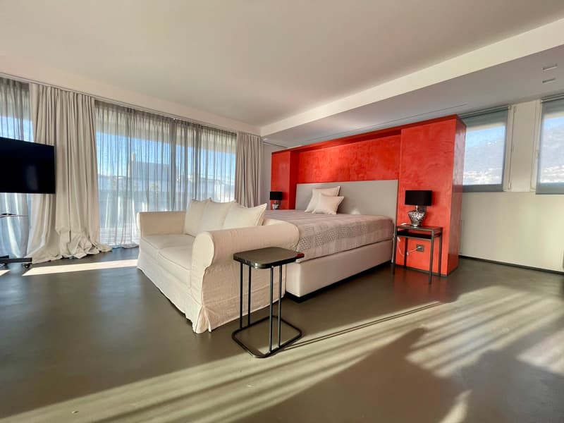 modern white furniture, red wall, bed with pillows, couch with pillows, side tables with lamps, TV, and large windows with curtains