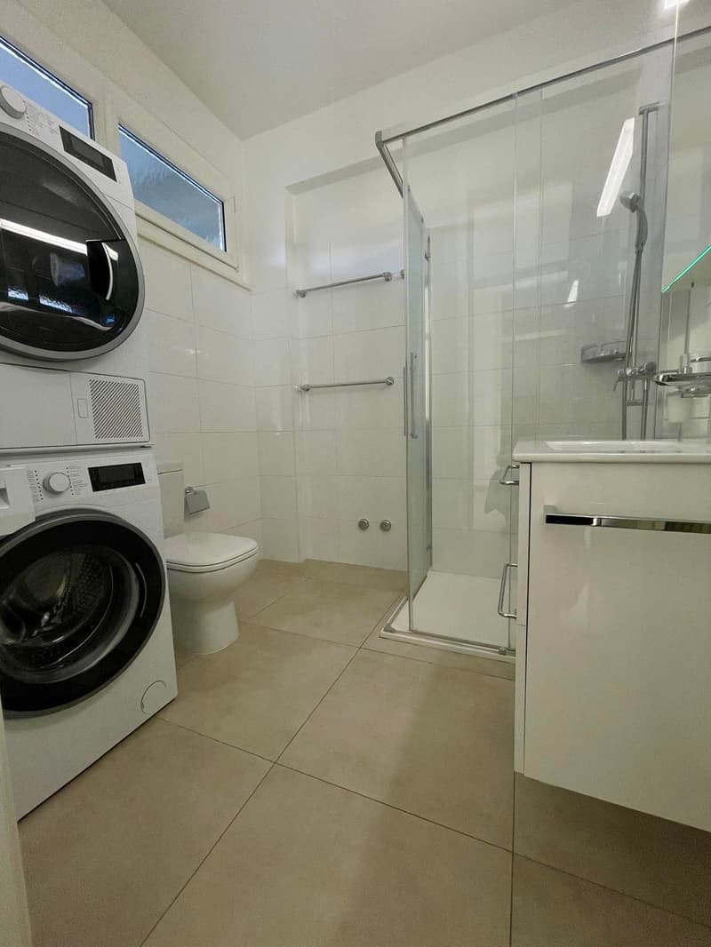 A modern, clean bathroom with a glass shower stall, white tiles, and tiled flooring. The room features a white washing machine and dryer stacked on top of each other, a white toilet, and a sink with a vanity.