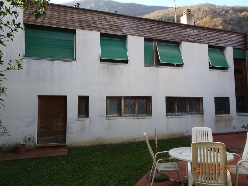 Large white building, wooden door, green shutters, tiled courtyard, outdoor seating, potted plant