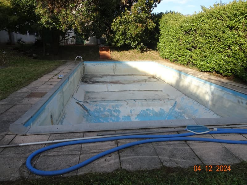 Empty swimming pool with a pool skimmer and a hose, surrounded by bushes