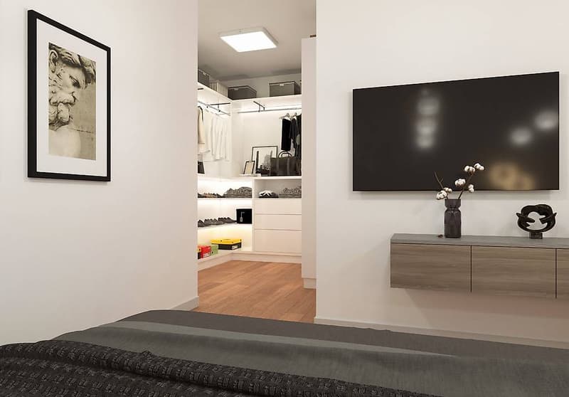 A bedroom with white walls and wooden flooring. There is a bed on the left, a television mounted on the wall, and a shelf on the opposite wall. The room also has a walk-in closet and a wardrobe. The shelf has decorative items such as a vase with flowers,