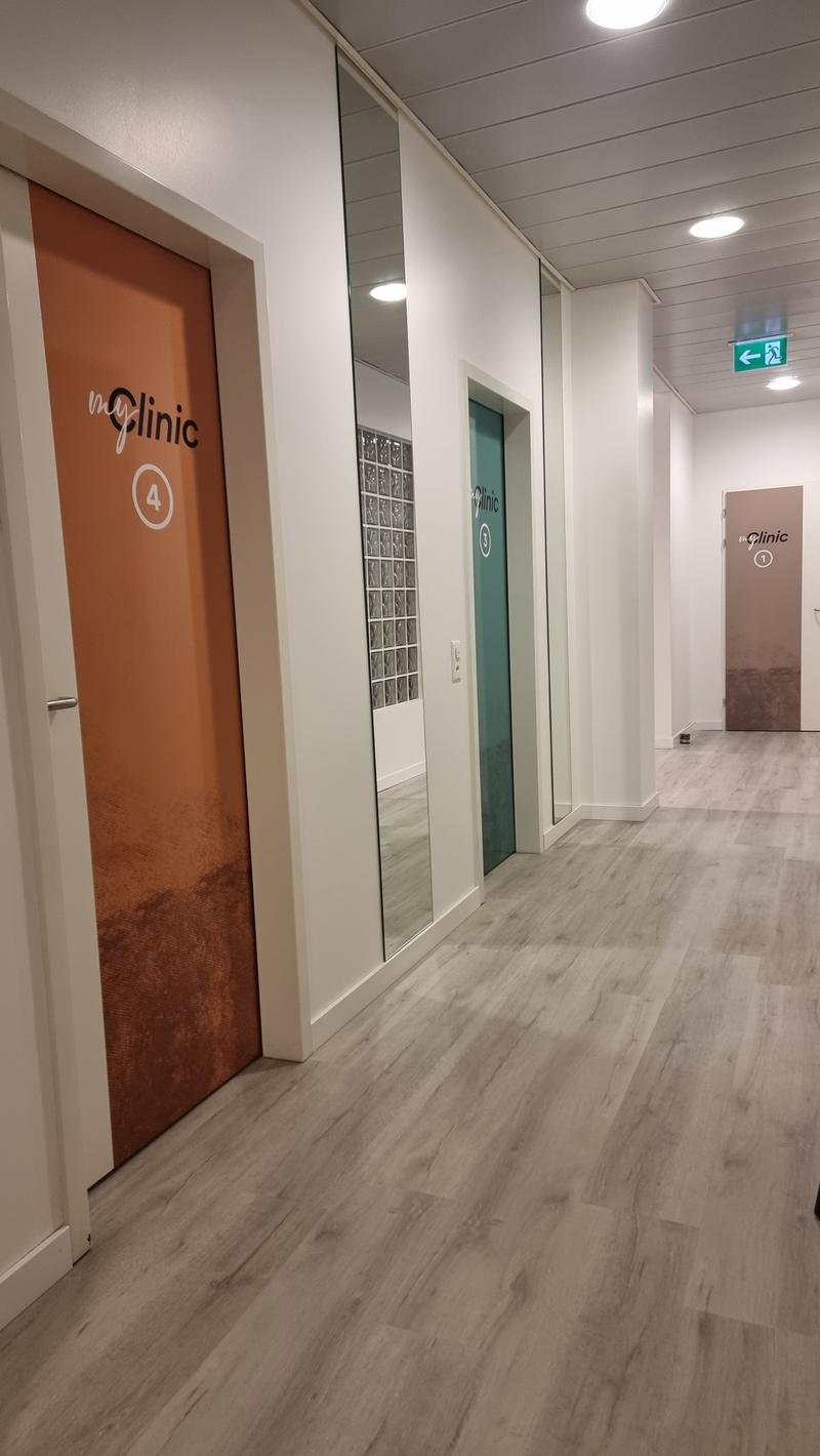 White walls, wooden floors, two doors with clinic signs and number, mirror, open space, empty