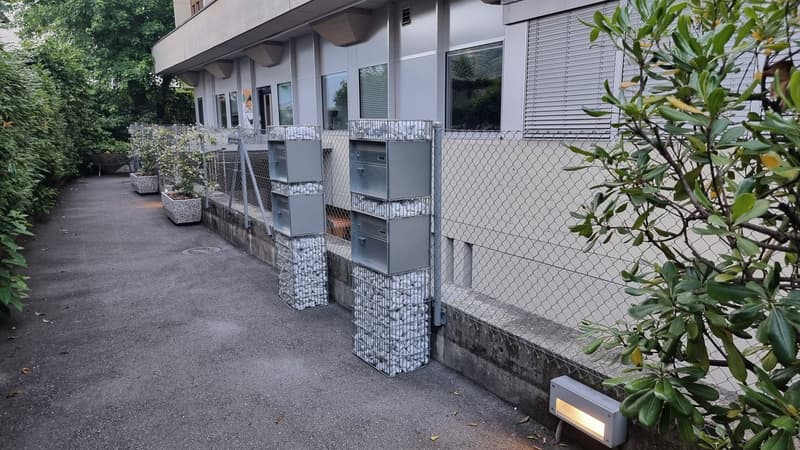 Building with multiple mailboxes along the front sidewalk, surrounded by a chain-link fence, with stone planters, potted plants, and trees.