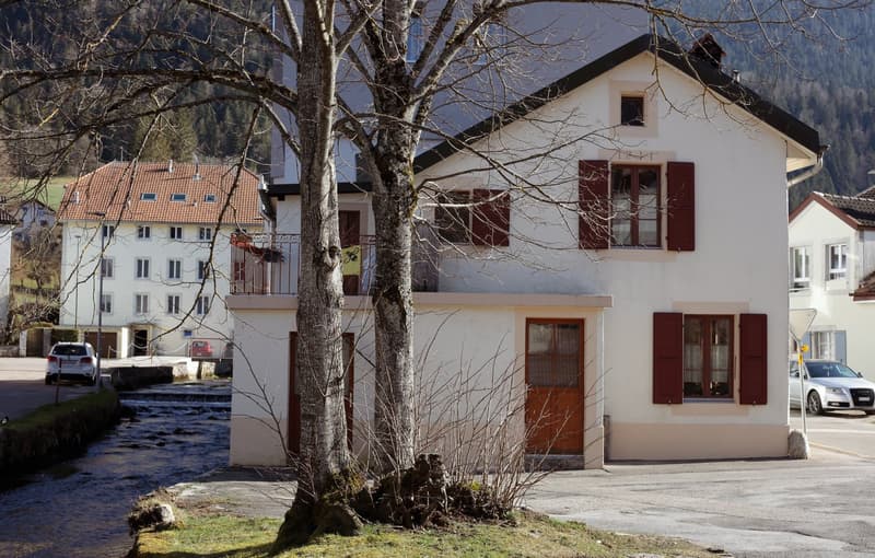 white, raised ground floor, brown windows and doors, balcony, red roof, located in a village, near a stream