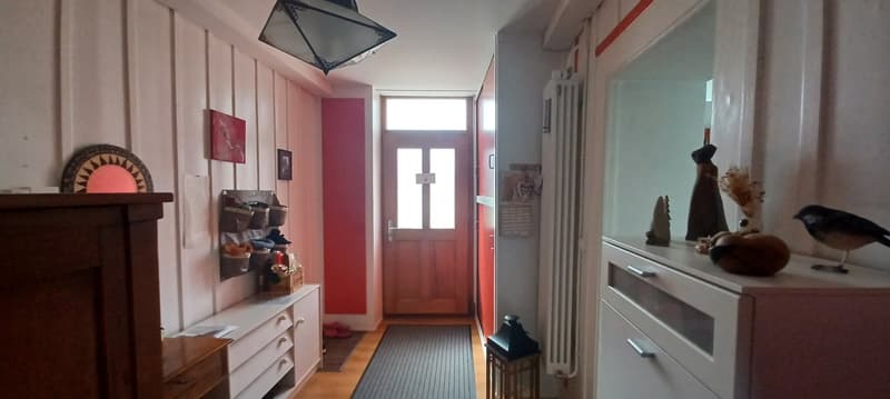 Decorated hallway with wooden floor, red painted wall, wooden door, shelves with baskets, picture frames, and a radiator. It has a hallway rug, and a lit lamp.