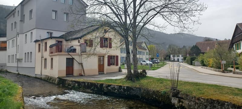a two story beige house with white windows, one balcony on the second floor, next to a river, with buildings and mountains in the background