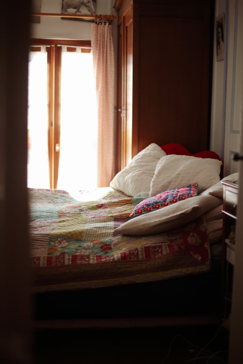 A bedroom with a bed and pillows, wooden doors, curtains, framed picture on wall, bright light from windows
