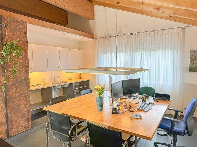 A room with a large wooden table, computer monitors, keyboard, mouse, calculator, scissors, pens, pencils, a vase with flowers, white curtains, wooden beam ceiling, kitchen with a sink, faucet, stove, and cabinets.