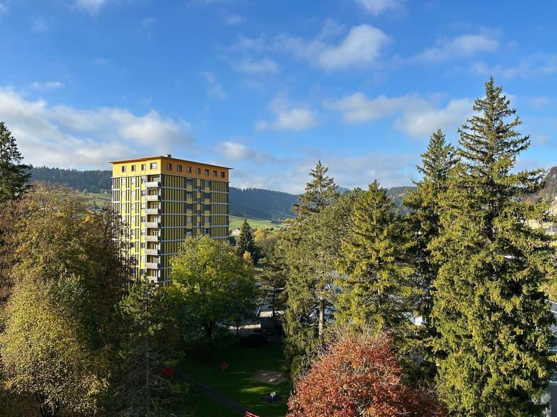 Apartment building with yellow and white color, many windows, surrounded by trees, hills in the background