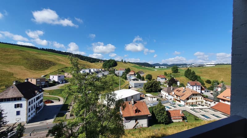 An aerial view of a hillside village with multiple buildings of different architectural styles, surrounded by lush green landscape, visible swimming pools and a building in the distance under a partly cloudy sky