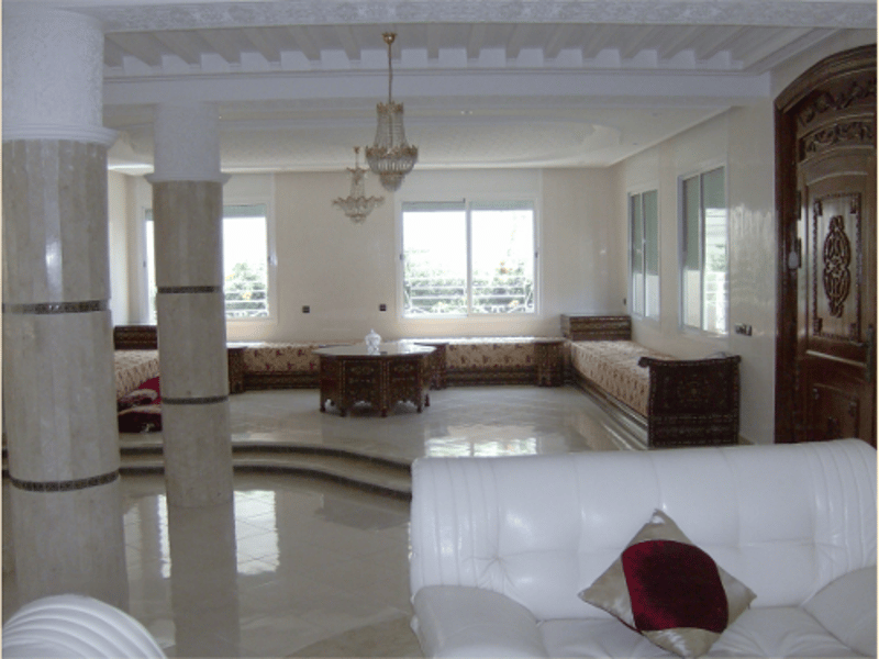 White marble floor, decorative columns, arched doorway, windows with railings, white leather couch, table, cushioned seating, chandeliers