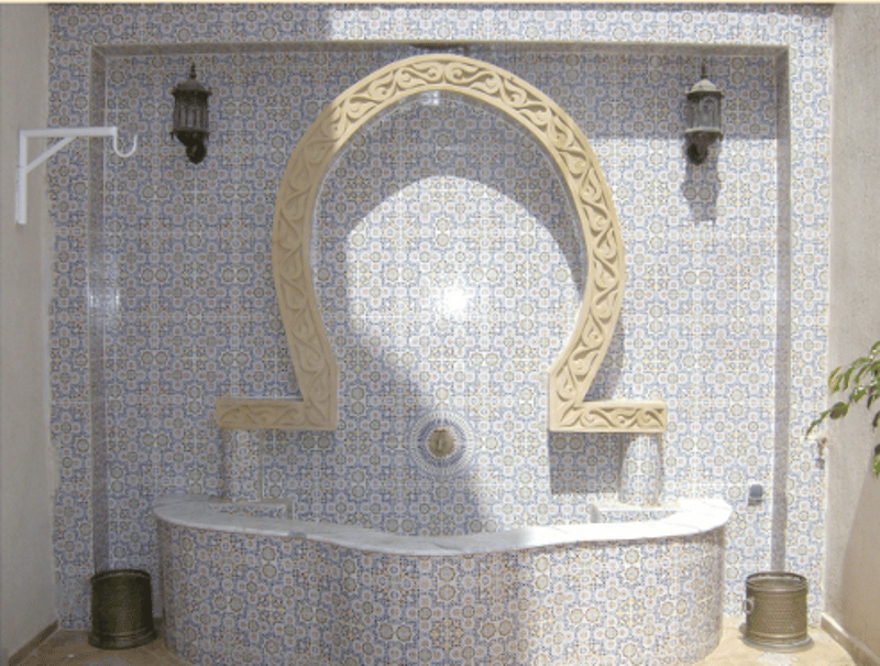 An ornate bathroom with blue tiles, double sinks, and a curved stone structure with a central fountain, flanked by decorative lamps.