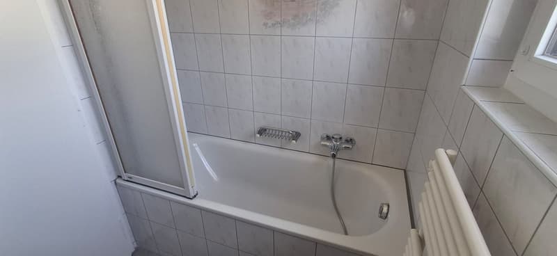 Bathroom: white tile walls, tile floor, white bathtub, glass shower door, shower faucet, and a white radiator.