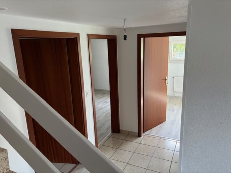 Empty room with white walls, tiled floor, wooden doors, and a staircase.