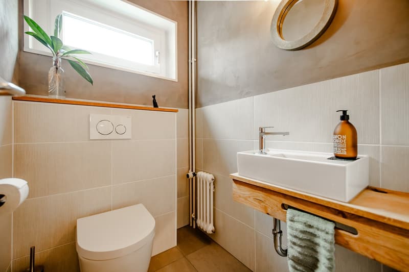 modern bathroom with white tiles, white toilet, white sink, wooden countertop, white radiator, plant on window sill, round mirror on wall