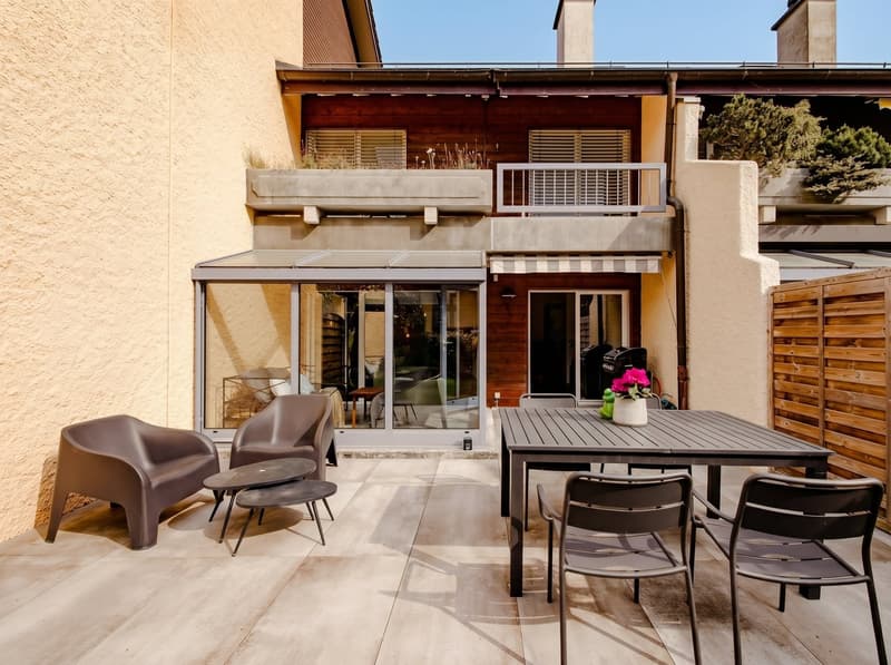 Outdoor patio with a table, chairs, and a small table with a flower vase, a wooden fence, and a glass wall.