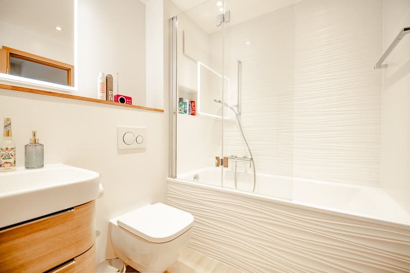Bathroom with white tiles, white bathtub, white sink, wooden cabinet, white toilet, shower, mirror, and shelves with toiletries