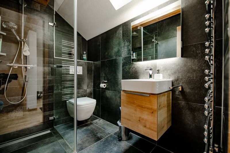 modern bathroom with glass shower, white toilet, wooden cabinet, white sink, mirror, and black tiles