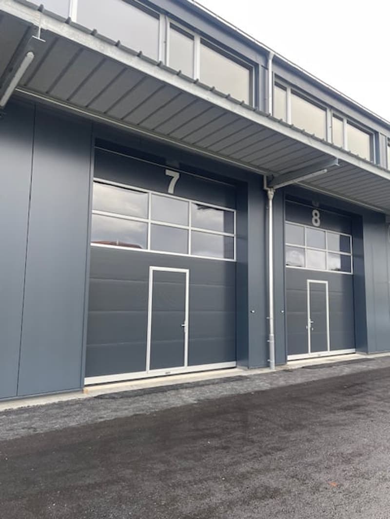 Gray facade with two garage doors, numbered 7 and 8, metal awning, and a large parking lot in front.