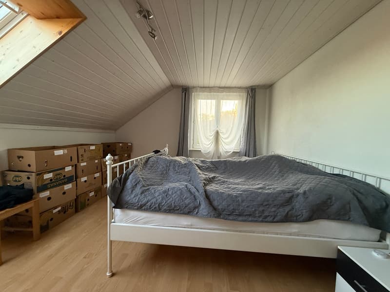 A bedroom with a bed, wooden floor, white walls, a window with curtains, and a ceiling with wooden beams and lights. There are also several storage boxes stacked on the left side of the room.