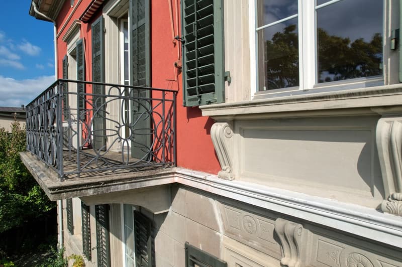 Balcony with black railing, red wall, green shutters, white windows, decorative elements