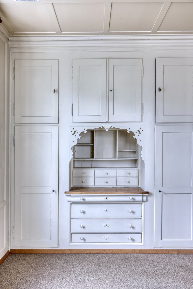 A room with white wooden cabinets, drawers, and shelves. The room has a wooden floor and a white ceiling.