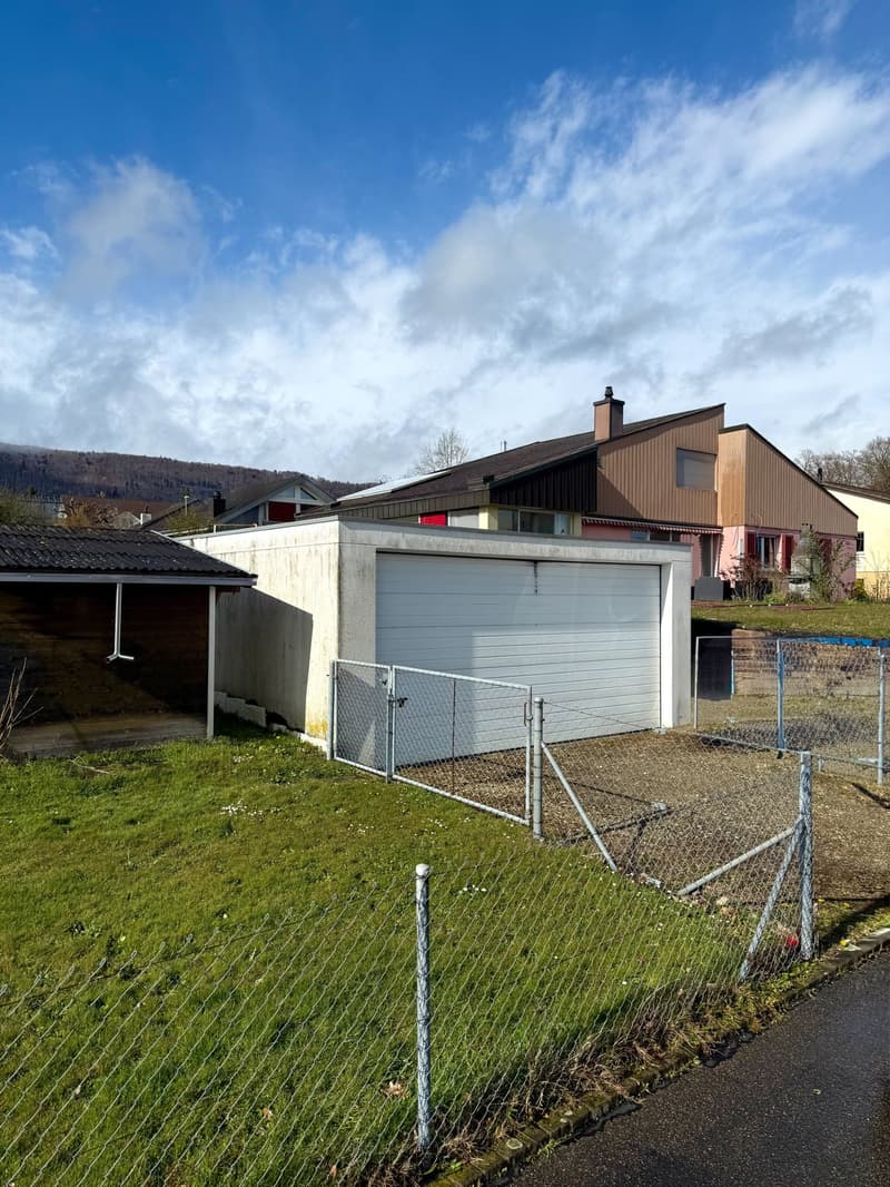 Detached house with a garage in front, surrounded by a fence, with a grassy area and a mountain in the background.